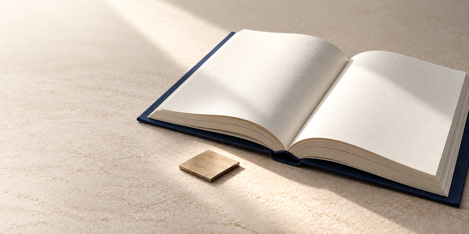 An open navy-covered book with blank parchment pages sits on pale limestone, with a single gold square in front.
